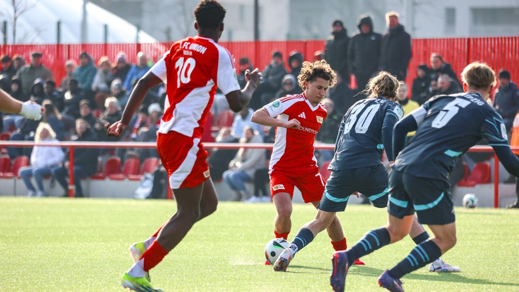 Spielszene eines Fußballspiels mit Spielern in roten und grauen Trikots auf einem Kunstrasenplatz, Zuschauern im Hintergrund.