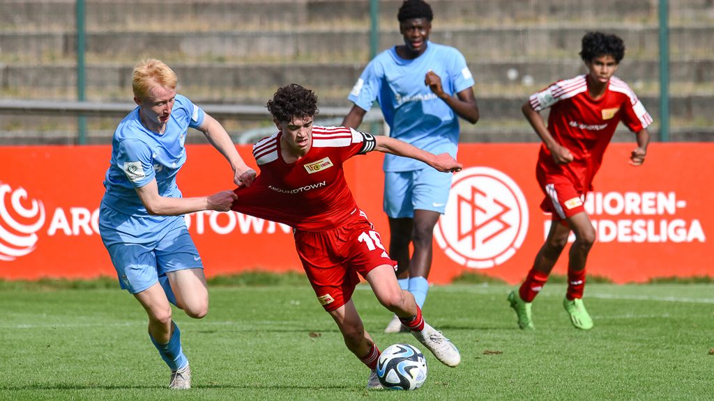 Junger Fußballspieler mit Kapitänsbinde dribbelt den Ball, während ein Gegner ihn am Trikot festhält. Im Hintergrund stehen weitere Spieler.