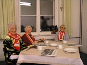 Drei ältere Frauen sitzen an einem Tisch mit gedecktem Geschirr und lächeln in die Kamera, jeweils mit Schals in den Farben Rot und Weiß.