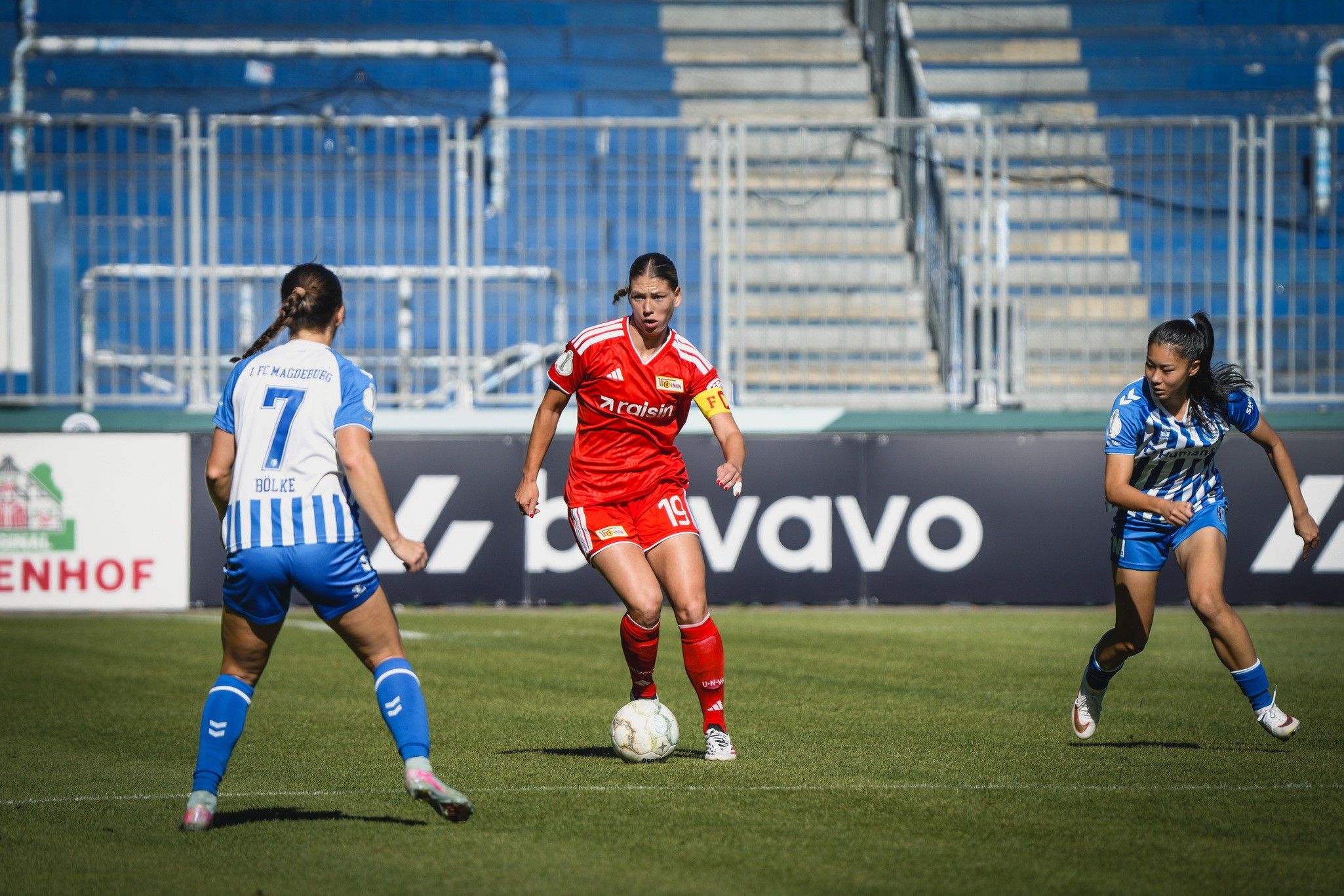 Eine Fußballspielerin in roter Uniform dribbelt den Ball, während zwei Spielerinnen in blauen Trikots sie pressen.