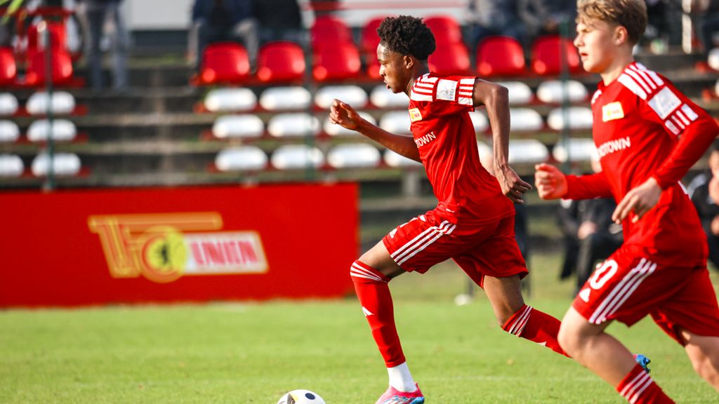 Zwei junge Fußballspieler in roten Trikots rennen über das Spielfeld, während Zuschauer im Hintergrund stehen.