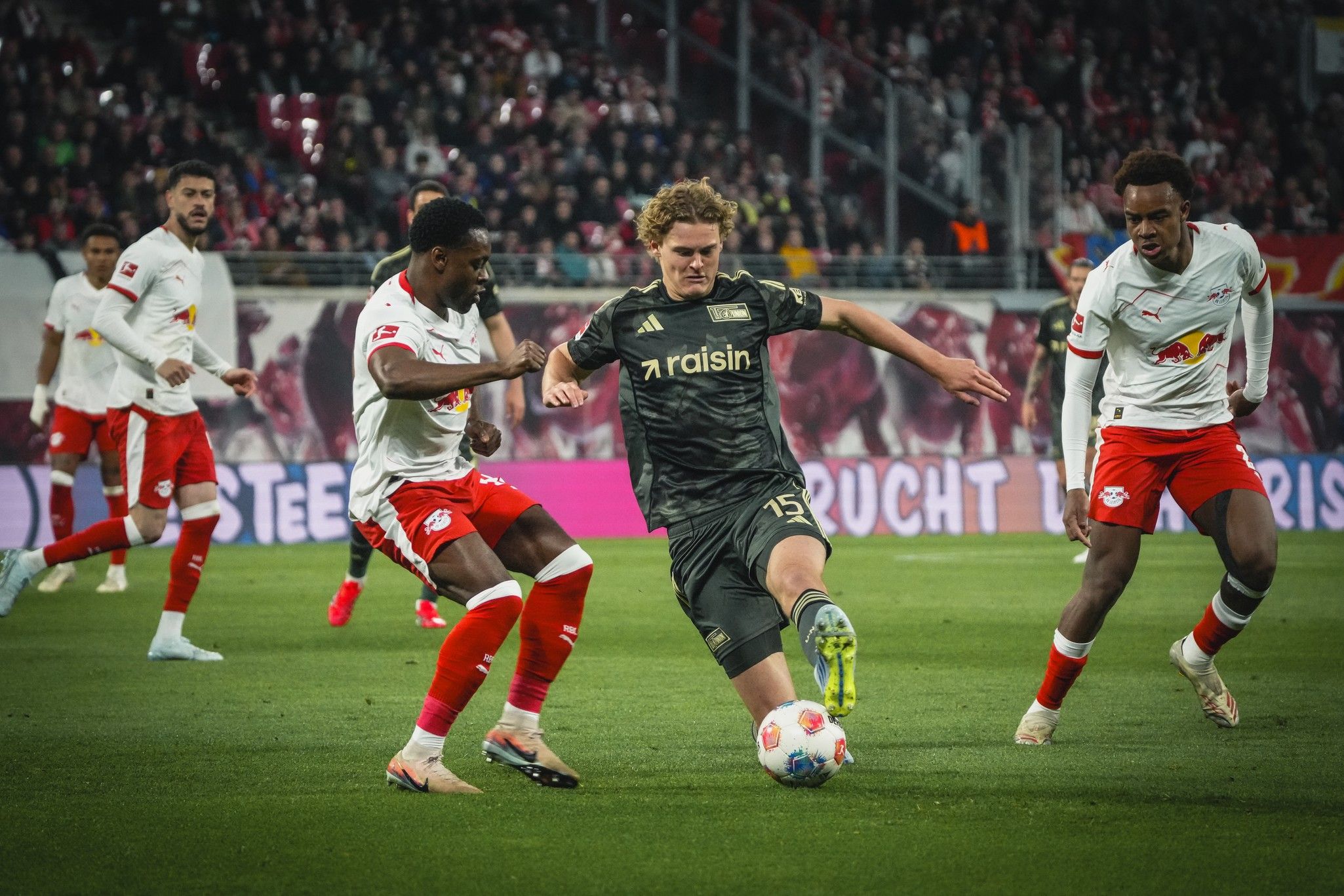 Ein Fußballspieler mit blondem Haar dribbelt den Ball zwischen zwei gegnerischen Spielern in einem Stadion.