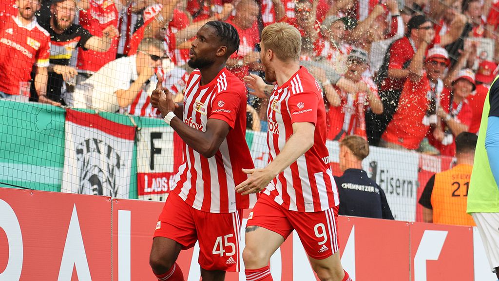 Two soccer players in red-and-white jerseys celebrate a goal in front of a cheering crowd in the stadium.