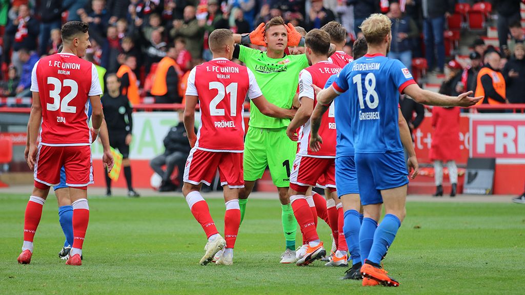 Eine Gruppe von Fußballspielern in roten und blauen Trikots steht auf dem Spielfeld, während der Torwart sich den Kopf hält.