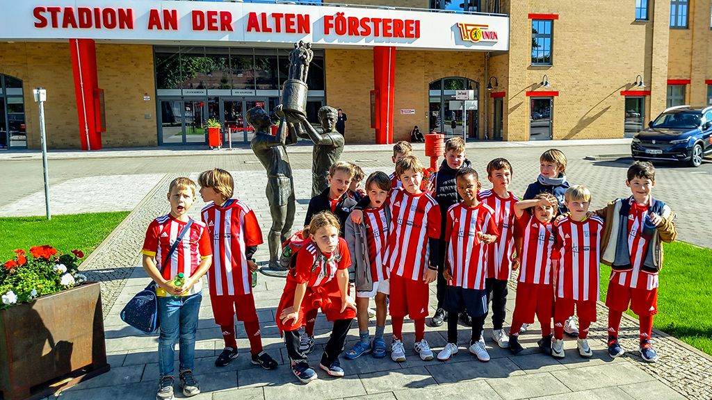 Gruppe von Kindern in rot-weißen Trikots steht vor dem Stadion an der alten Försterei, neben einer Statue.