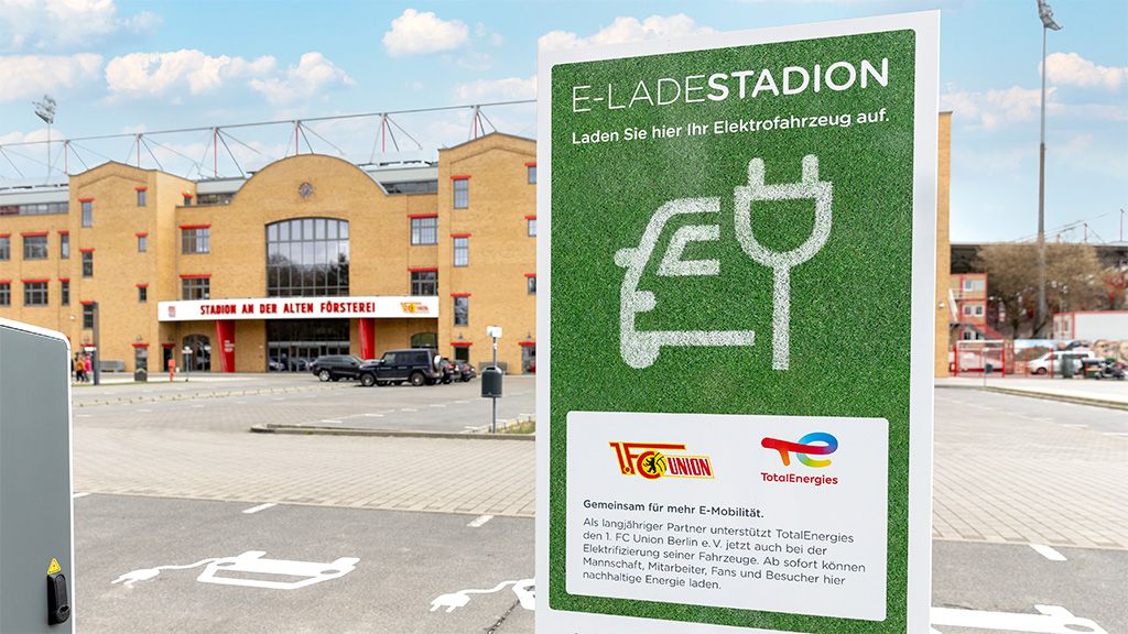 Ein Parkplatz mit einem Schild für E-Ladestationen vor einem Stadion mit rot-weißer Fassade.