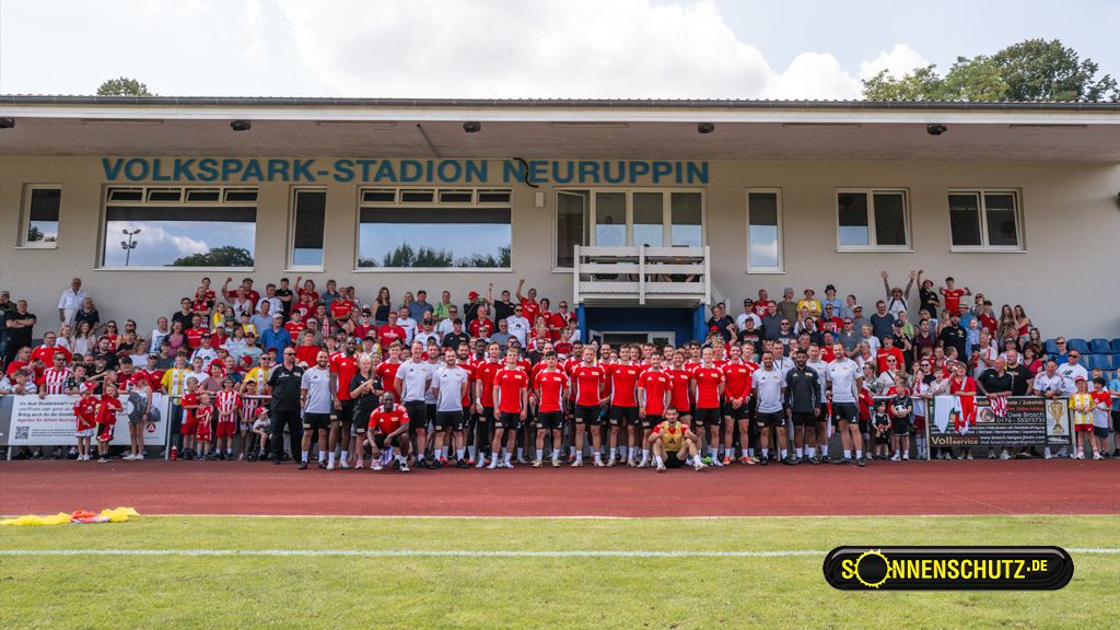 Gruppenshot von Fußballspielern und Fans im Volkspark-Stadion Neuruppin, umgeben von Zuschauern und Teammitglieder in Trikots.
