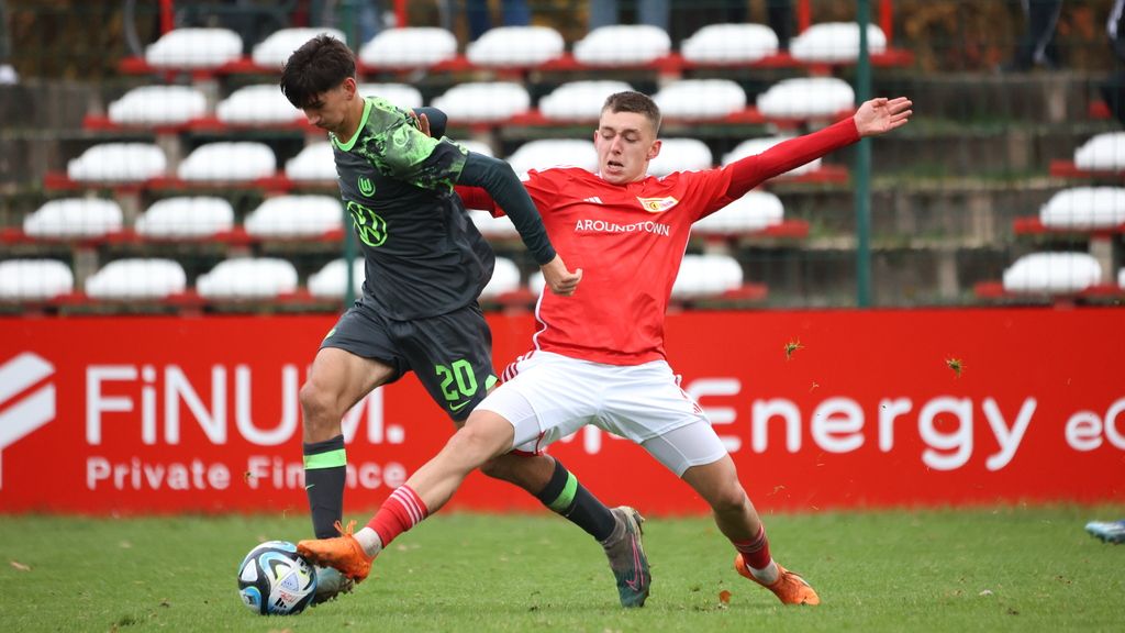 Ein Fußballspieler in grüner Trikotfarbe kämpft um den Ball gegen einen Spieler in rotem Trikot auf einem Rasenfeld.
