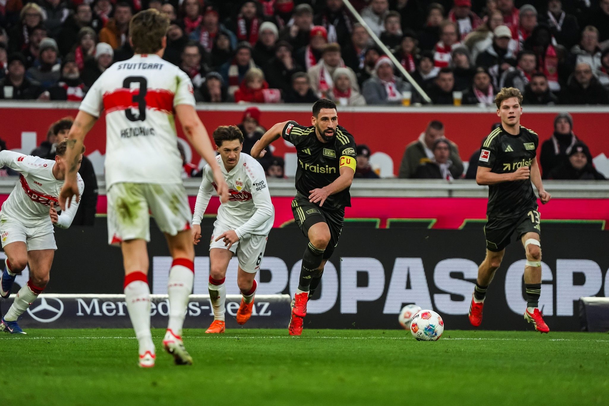 Fußballspielszene mit Spielern in schwarzen und weißen Trikots auf dem Spielfeld, Zuschauer im Hintergrund.