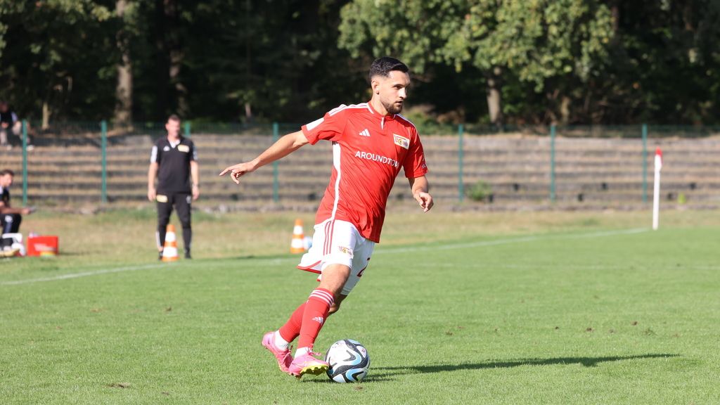 Ein Fußballspieler in rotem Trikot steht auf einem grünen Rasenfeld und bereitet sich auf einen Pass oder Schuss vor.