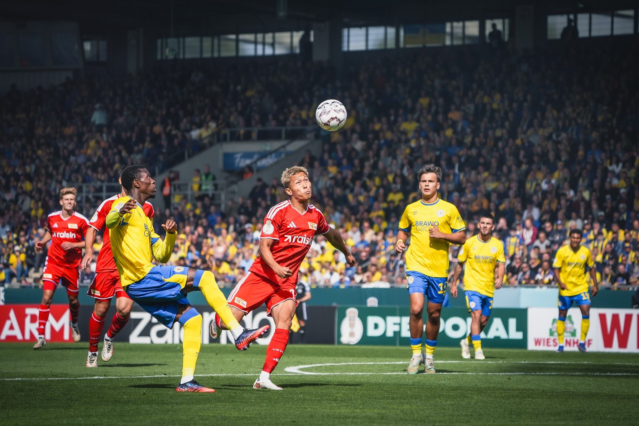 Spielszene im Fußball mit Spielern in roten und gelben Trikots, ein Spieler springt hoch, um den Ball zu erreichen, Publikum im Hintergrund.