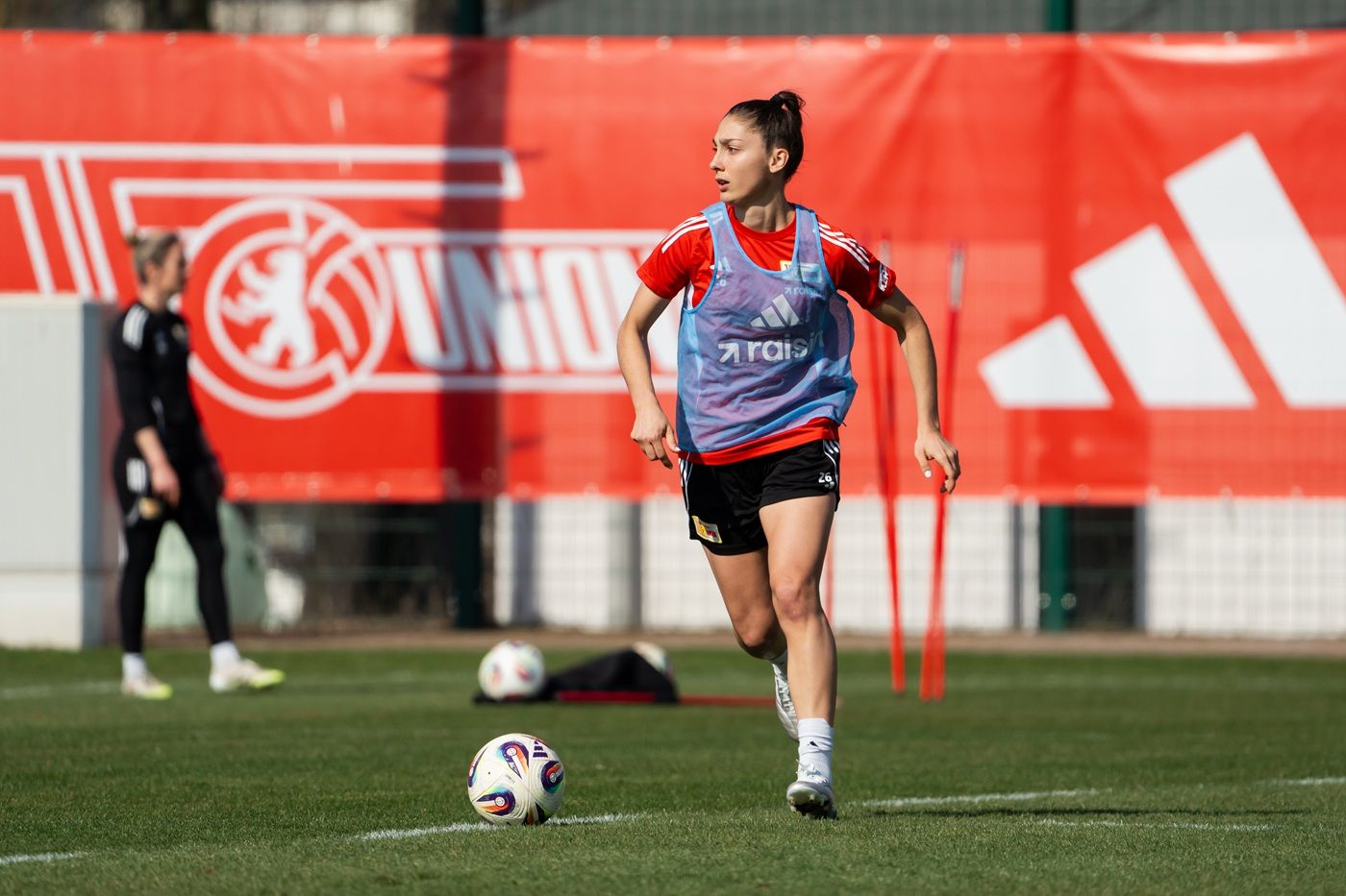 Eine sportliche Frau in Trainingskleidung läuft über ein Fußballfeld, während im Hintergrund ein rotes Banner sichtbar ist.