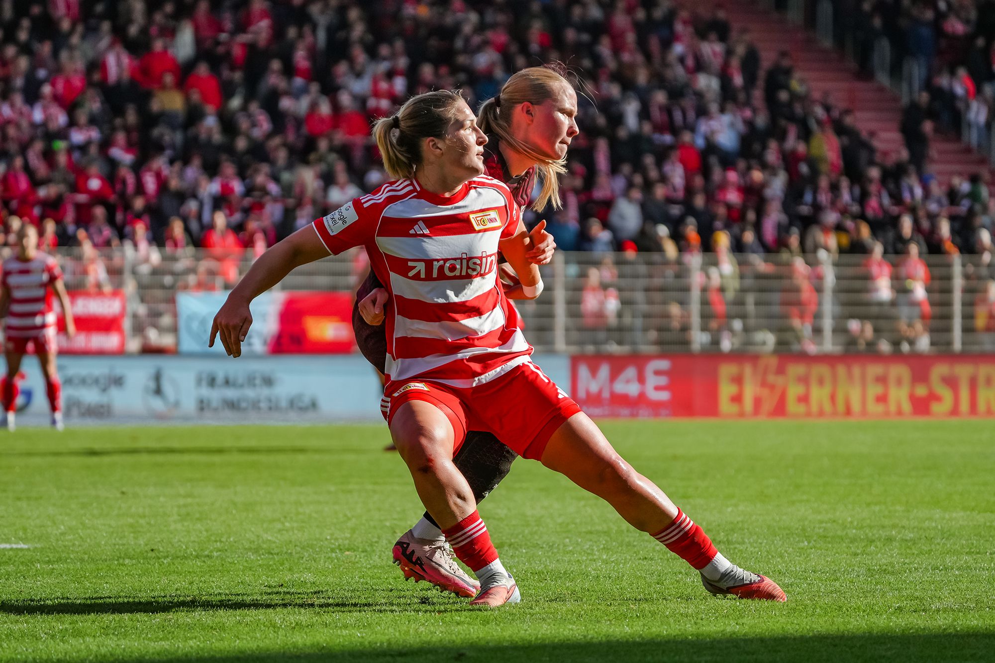Eine Spielerin in gestreiftem Trikot kämpft um den Ball in einem Fußballspiel auf dem Spielfeld vor jubelndem Publikum.