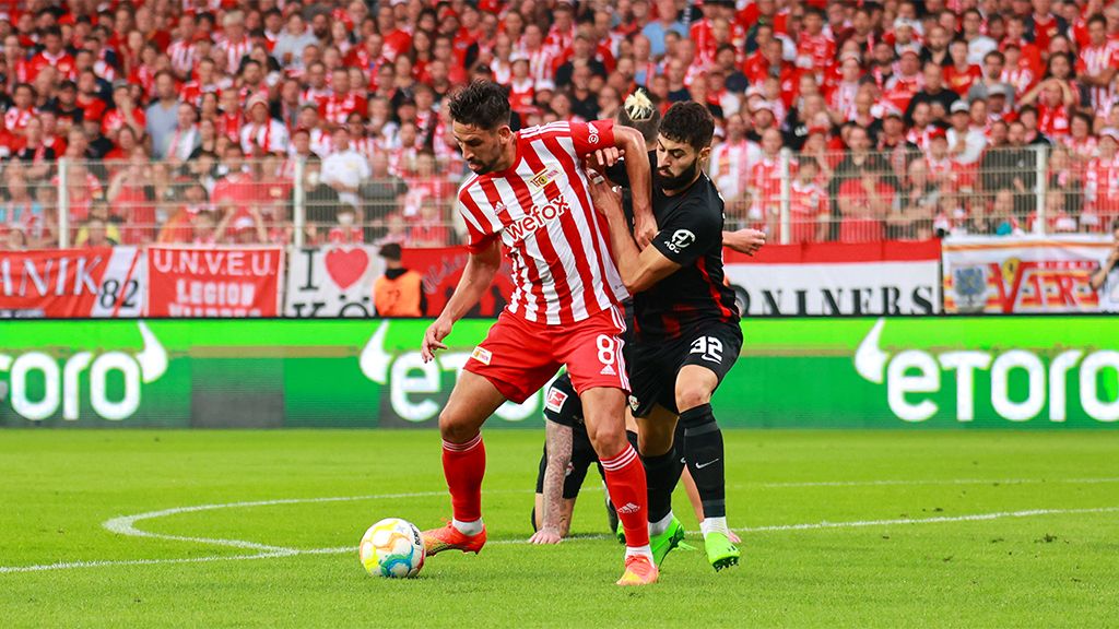 Ein Fußballspieler in rot-weißer Kleidung dribbelt den Ball, während ein gegnerischer Spieler ihn von hinten herausfordert.