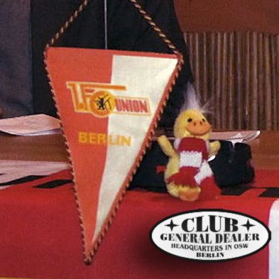 Wimpel des FC Union Berlin mit einem kleinen Plüschvogel in Vereinsfarben, auf einem Tisch liegend.
