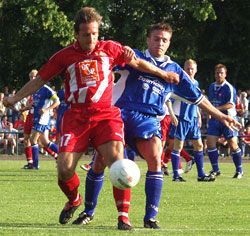 Zwei Fußballspieler kämpfen um den Ball auf einem Rasenfeld, während Zuschauer im Hintergrund stehen.