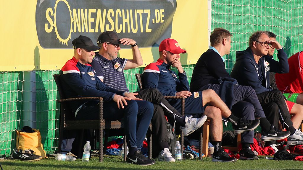 Fünf Personen sitzen auf einer Bank neben einem Fußballfeld und schauen nachdenklich auf das Spiel.