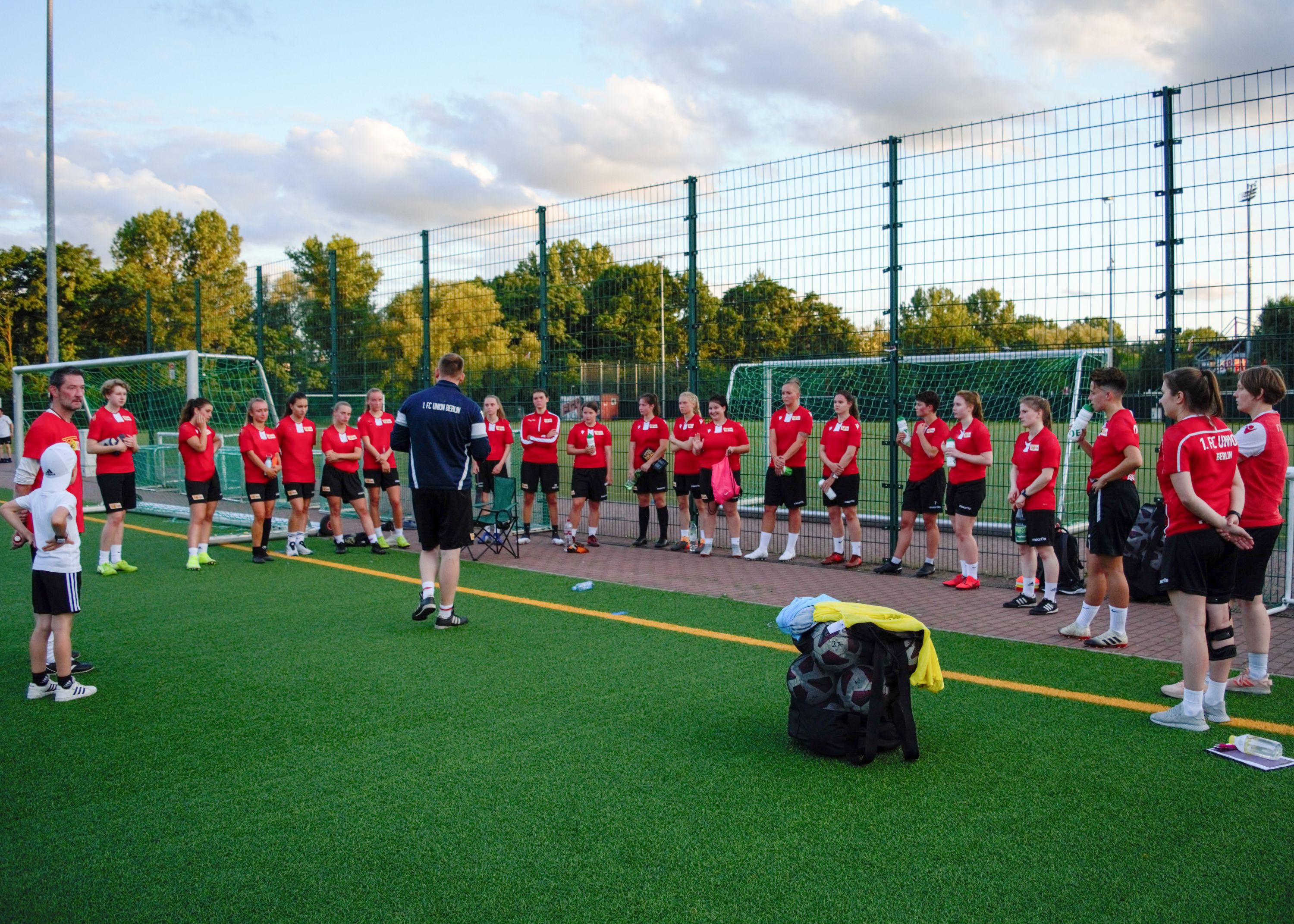 Eine Gruppe von Sportlern in roten Shirts steht im Kreis auf einem Trainingsplatz und hört einem Trainer zu.