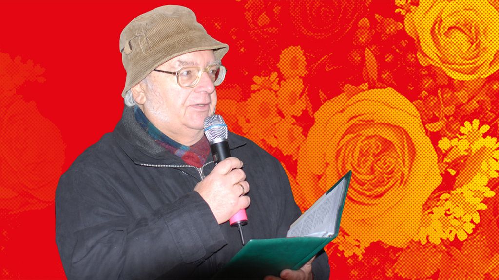 Ein Mann mit Brille und einem Braunen Hut hält ein Mikrofon und steht vor einem bunten, floralen Hintergrund.