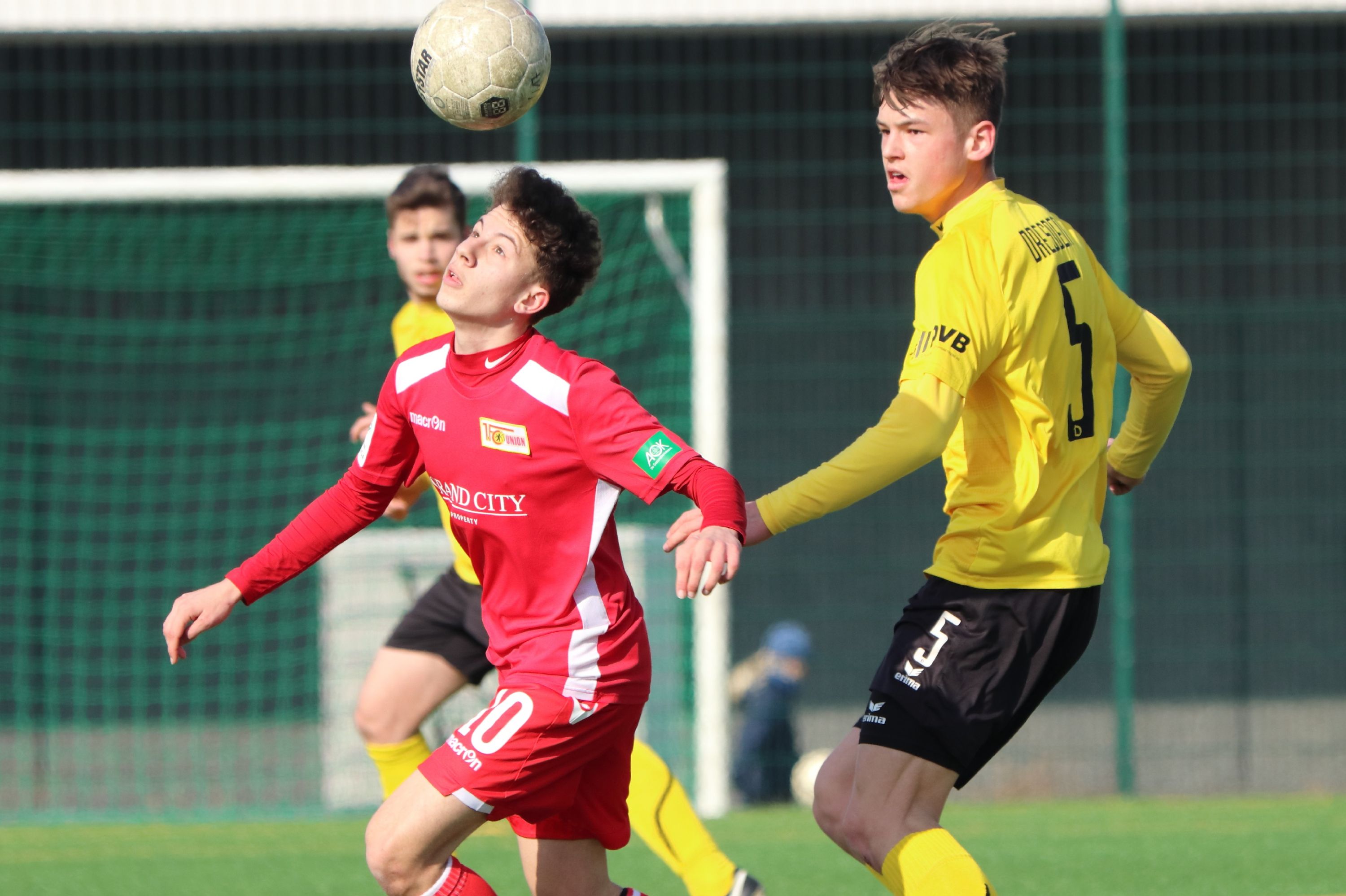 Two football players are fighting for the ball. One wears a red jersey, the other a yellow one. A goal is visible in the background.