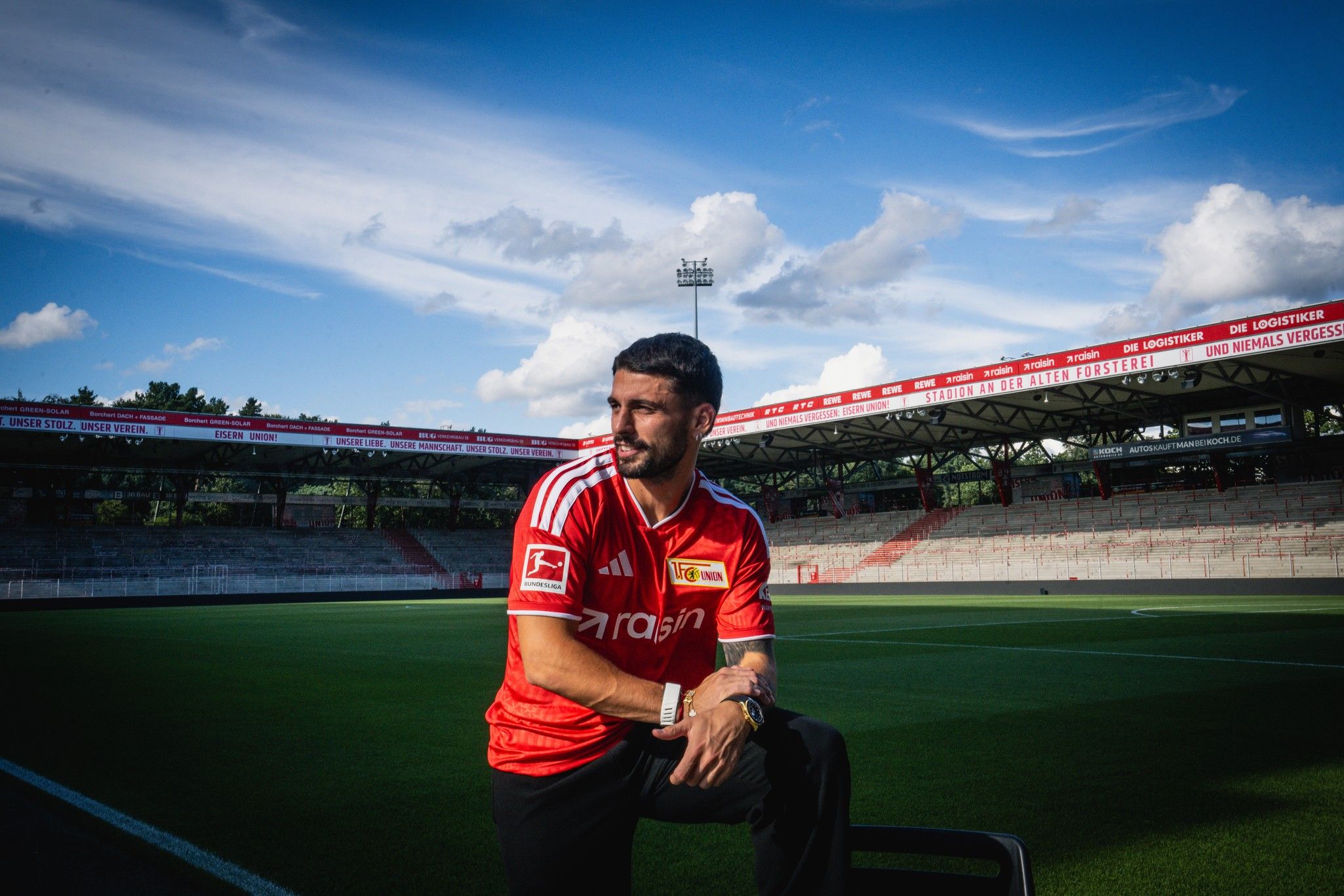 Ein Fußballspieler in einem roten Trikot sitzt auf einer Bank im leeren Stadion unter blauem Himmel und weißen Wolken.