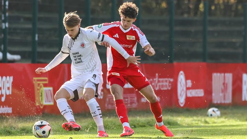 Zwei Fußballspieler in Aktion, einer in weißer, der andere in roter Uniform, auf einem Rasenplatz.