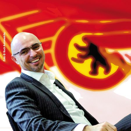 Mann in Anzug sitzt lächelnd vor einem farbigen Hintergrund mit einem stilisierten Logo.