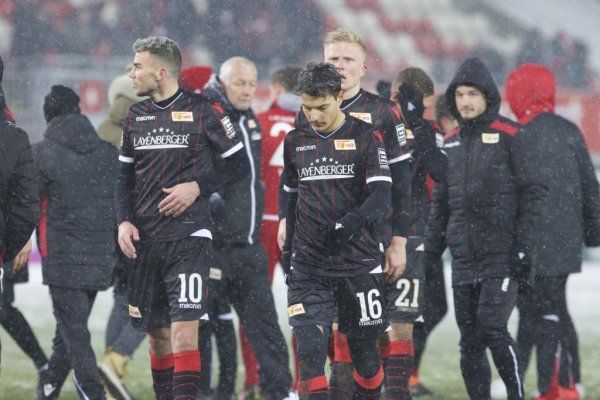 A football team in black jerseys leaves the field under snowfall, players look disappointed.