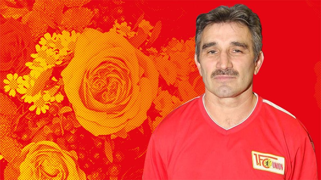 Mann mit einem Schnurrbart trägt ein rotes Shirt, vor einem Hintergrund mit gelben und roten Rosen.