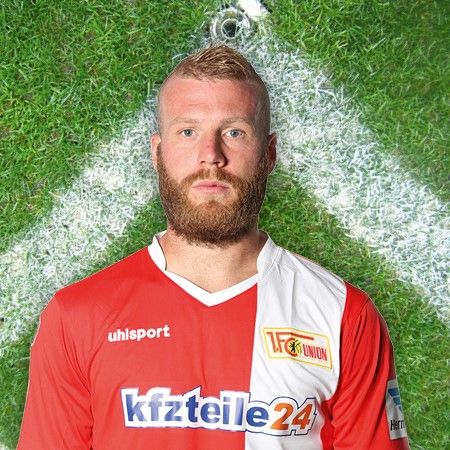 Ein Spieler des Fußballteams Union Berlin in rotem Trikot mit Logo, vor einem grünen Fußballfeld.