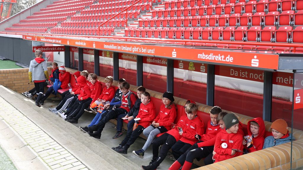Eine Gruppe von Kindern in roten Jacken sitzt auf einer Bank in einem Stadion, umgeben von leeren Tribünen.