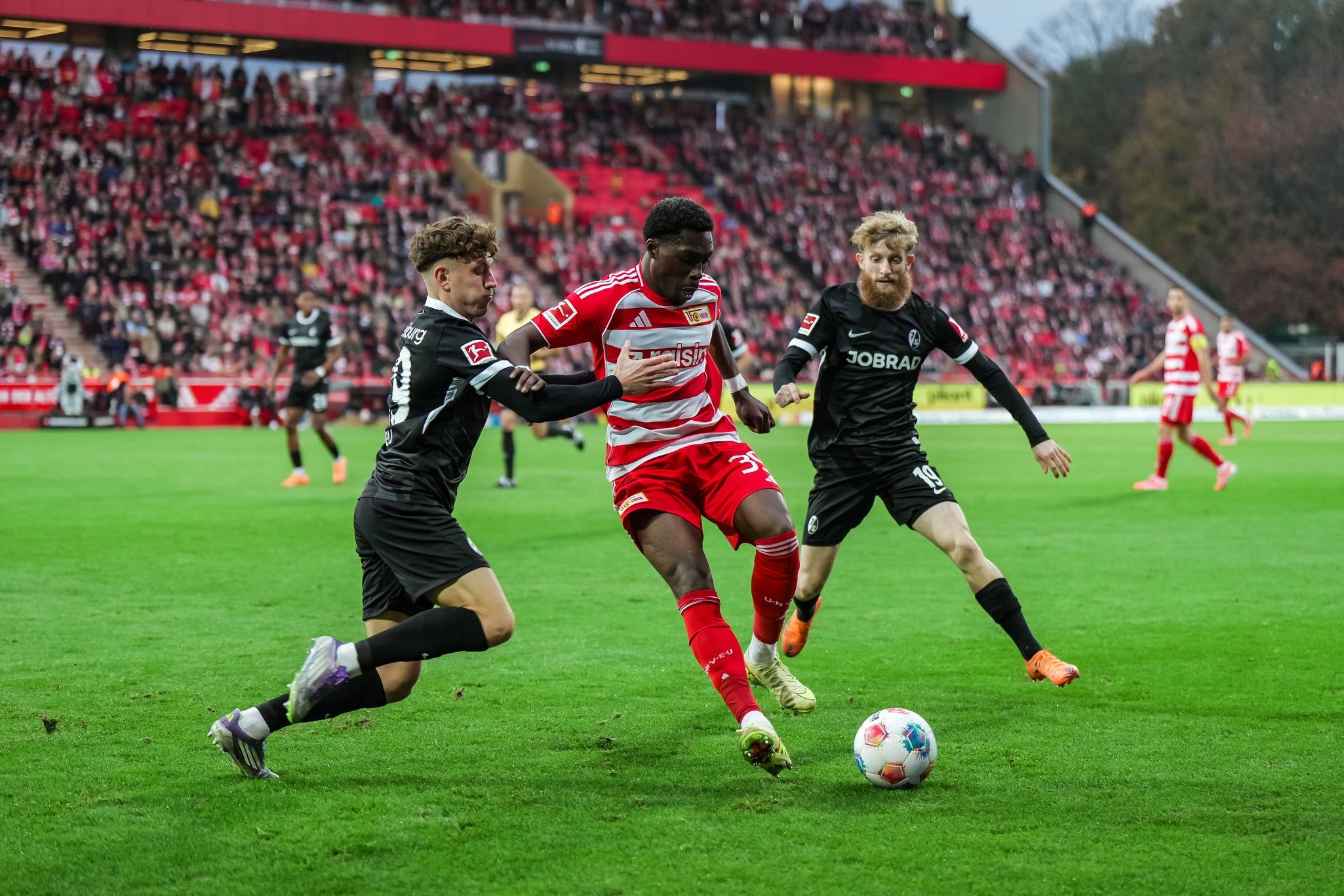 Spielszene im Fußballstadion: Ein Spieler in rot-weißem Trikot dribbelt den Ball, während zwei Gegner versuchen, ihn zu stoppen.