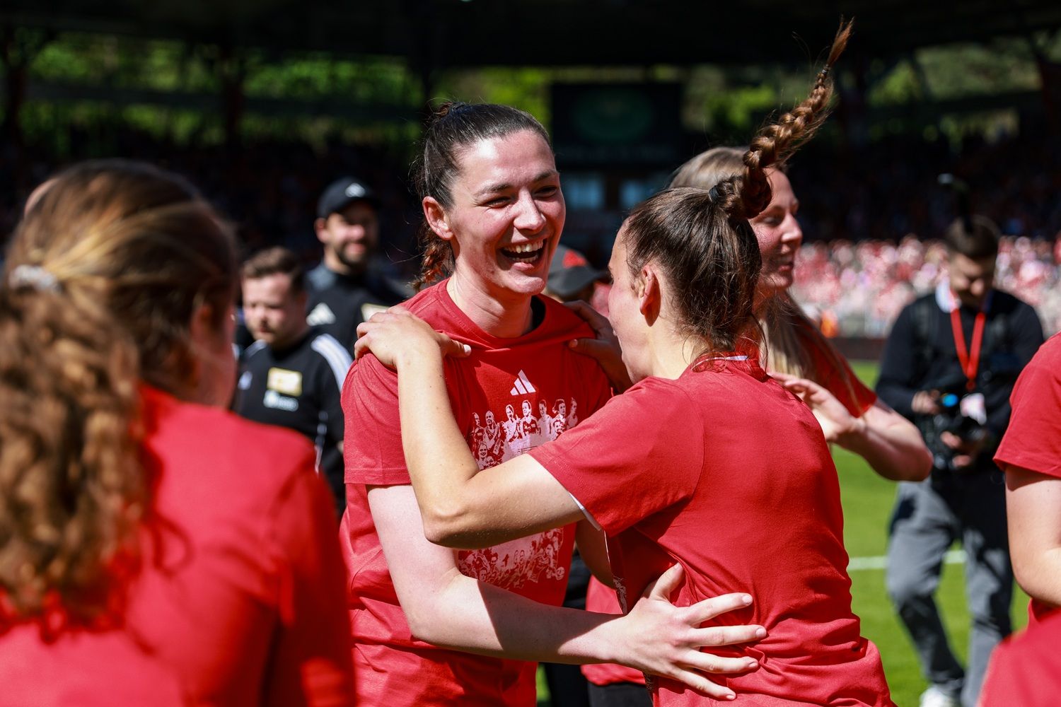 Zwei Fußballspielerinnen in roten Trikots umarmen sich lächelnd auf dem Spielfeld, umgeben von jubelnden Zuschauerinnen und Zuschauern.