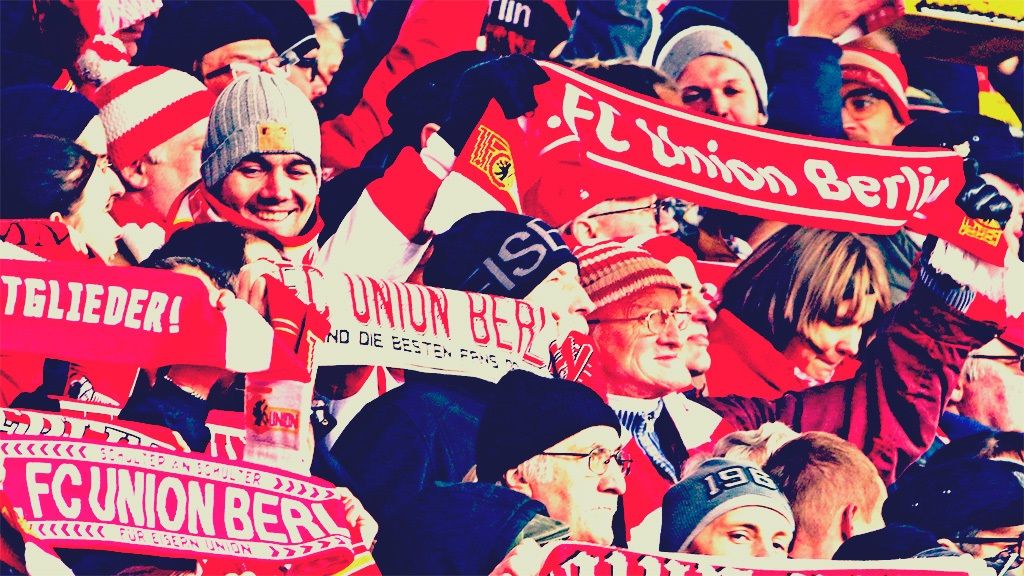 Fans von Union Berlin in roten und weißen Schals jubeln im Stadion und halten Banner hoch.
