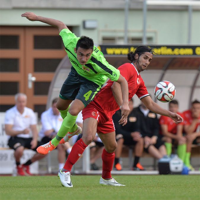 Zwei Fußballspieler im Wettkampf um den Ball, einer in grün, der andere in rot, während Zuschauer im Hintergrund beobachten.
