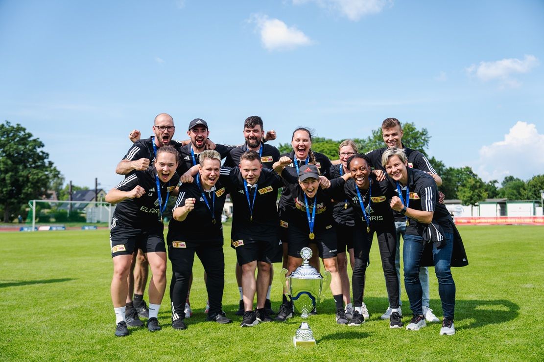 Eine Gruppe von Personen posiert auf einem Sportfeld, feiert mit einem Pokal und Medaillen unter blauem Himmel.