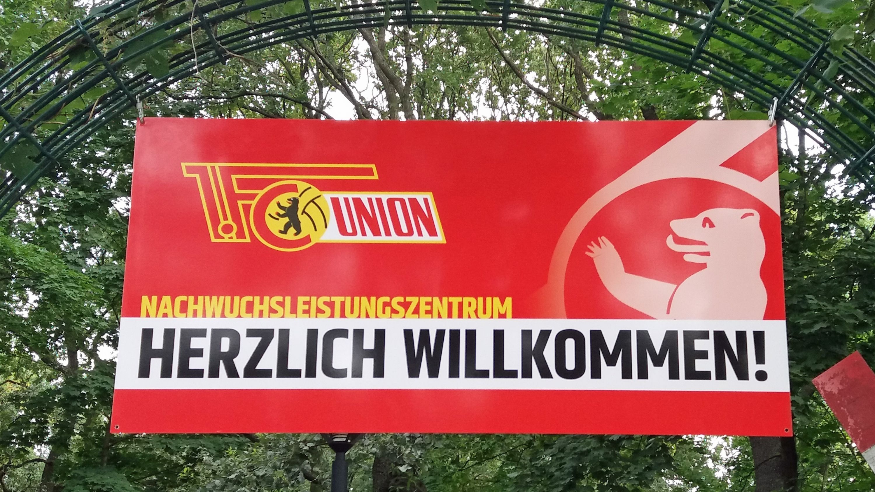 Banner mit der Aufschrift "Herzlich Willkommen!" und dem Logo des 1. FC Union. Umgeben von Bäumen und Pflanzen.
