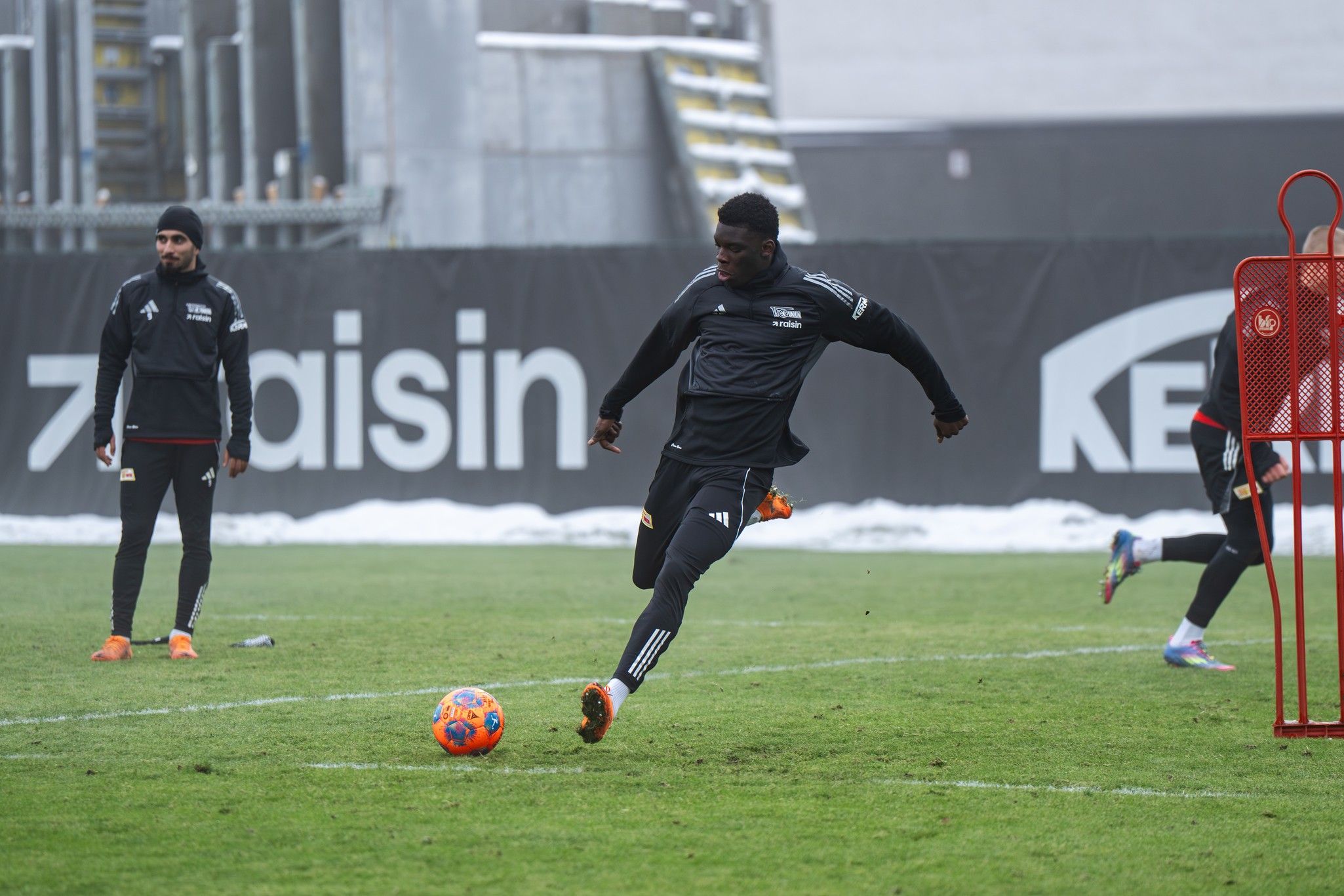 Ein Fußballspieler tritt einen Ball auf einem Trainingsplatz, während andere Spieler im Hintergrund zuschauen.