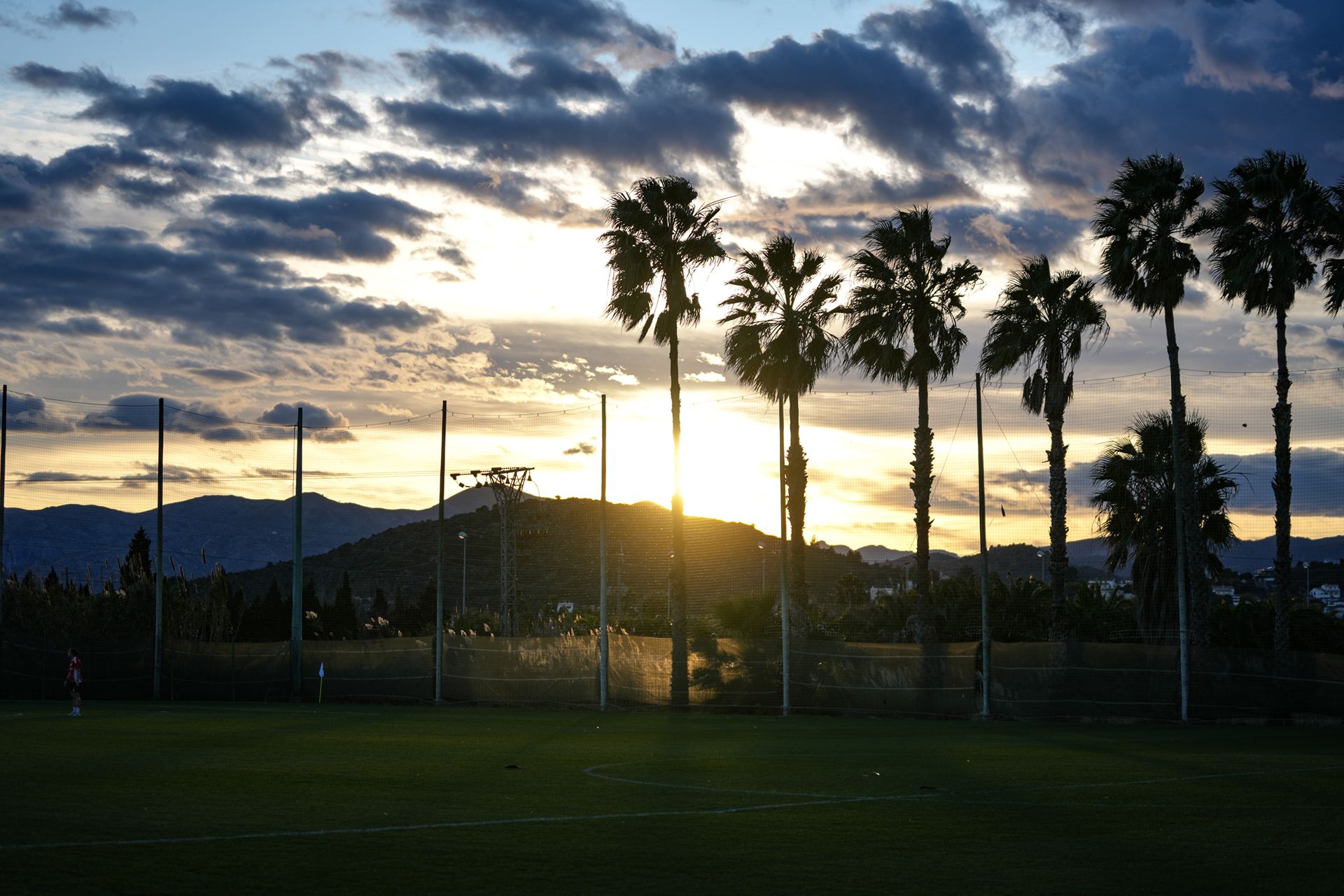Sonnenuntergang über einem Fußballfeld, umgeben von Palmen und Bergen im Hintergrund unter einem bewölkten Himmel.