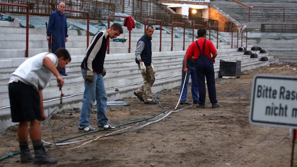 Arbeiter auf einer Baustelle in einem Stadion, die Kabel verlegen, mit einem Schild im Vordergrund, das auf Vorsicht hinweist.