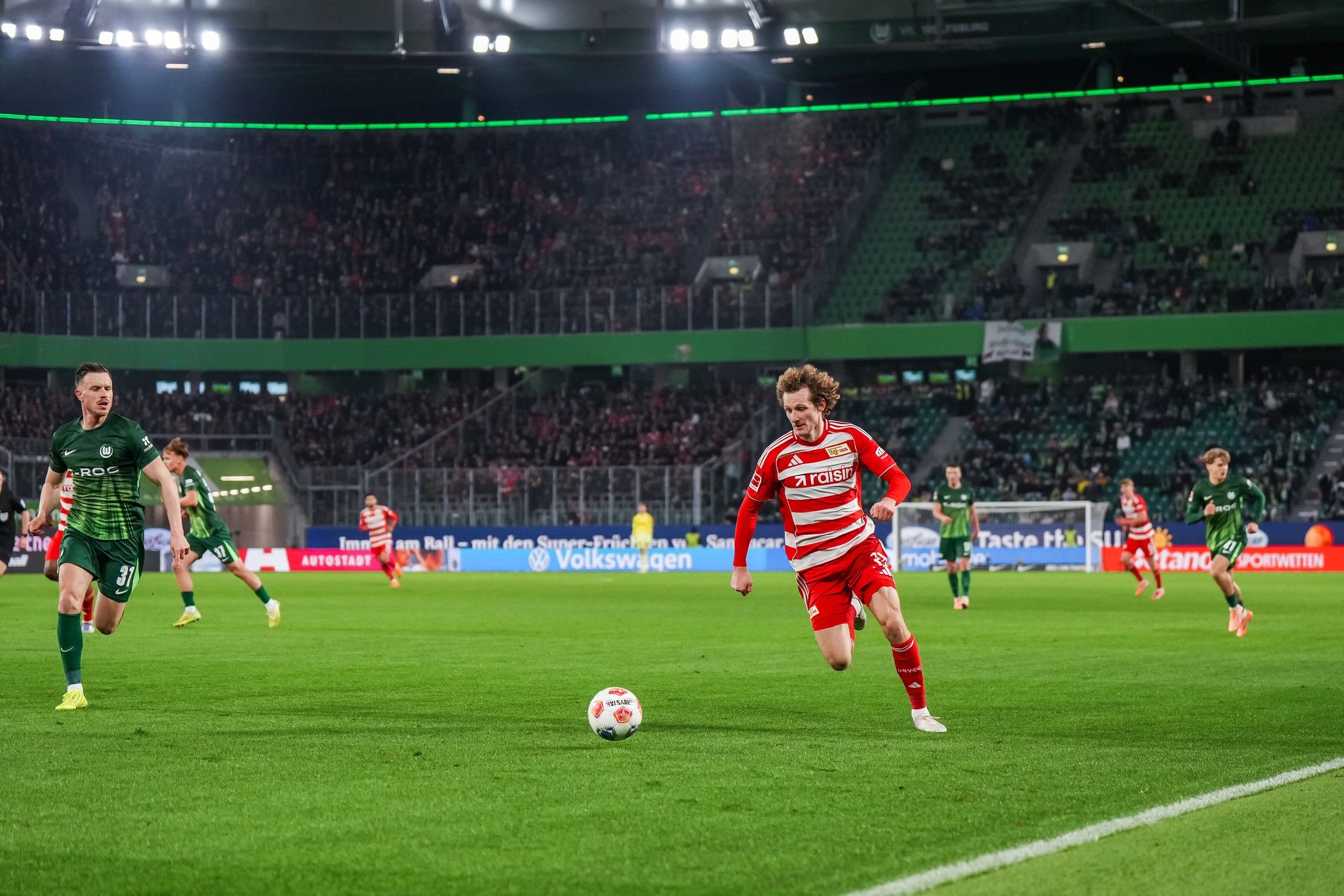 Spielszene im Fußball mit einem Spieler in rotem Trikot, der den Ball führt, während zwei Gegenspieler in grün folgen.