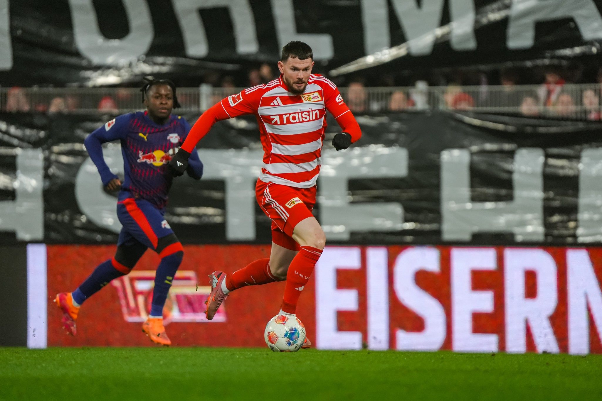 Ein Spieler in rotem Trikot mit weißen Streifen dribbelt den Ball, während ein Gegner in dunklem Trikot ihn verfolgt.