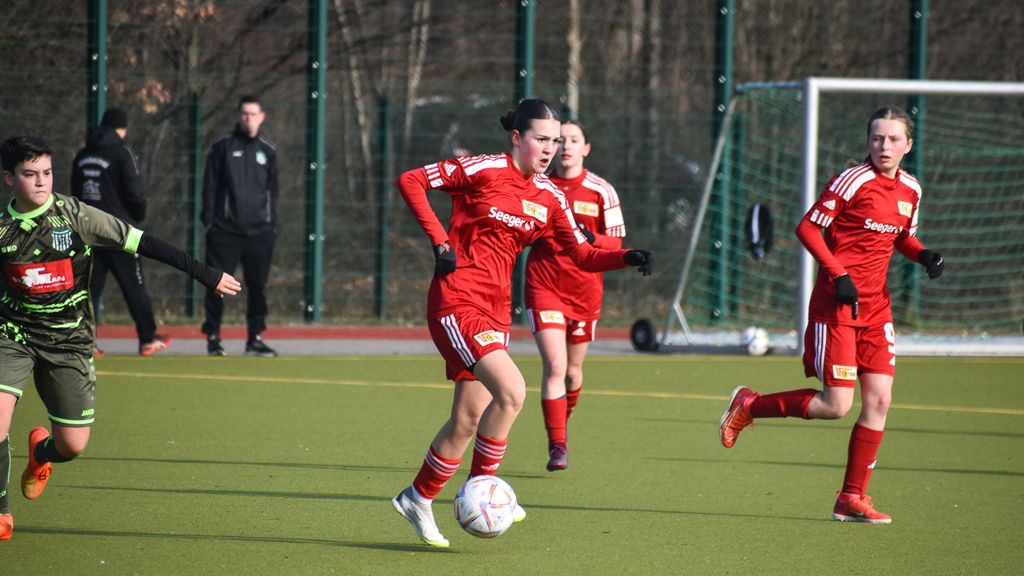 Eine Gruppe junger Frauen in roten Fußballtrikots spielt auf einem Feld, während eine Spielerin den Ball führt.