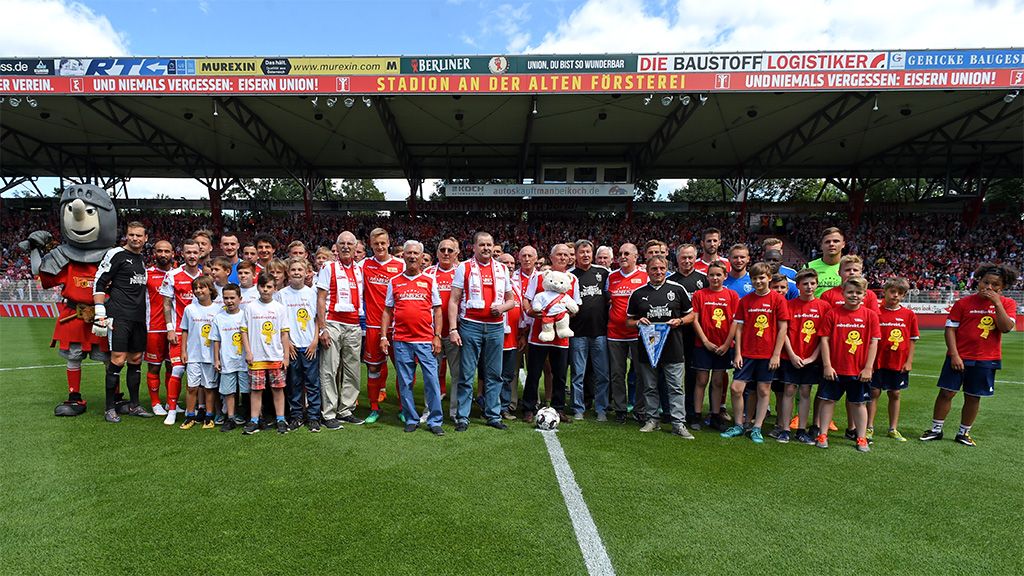 Gruppenszene mit Menschen in roten Trikots und T-Shirts auf einem Fußballfeld, umgeben von Zuschauern in einem Stadion.