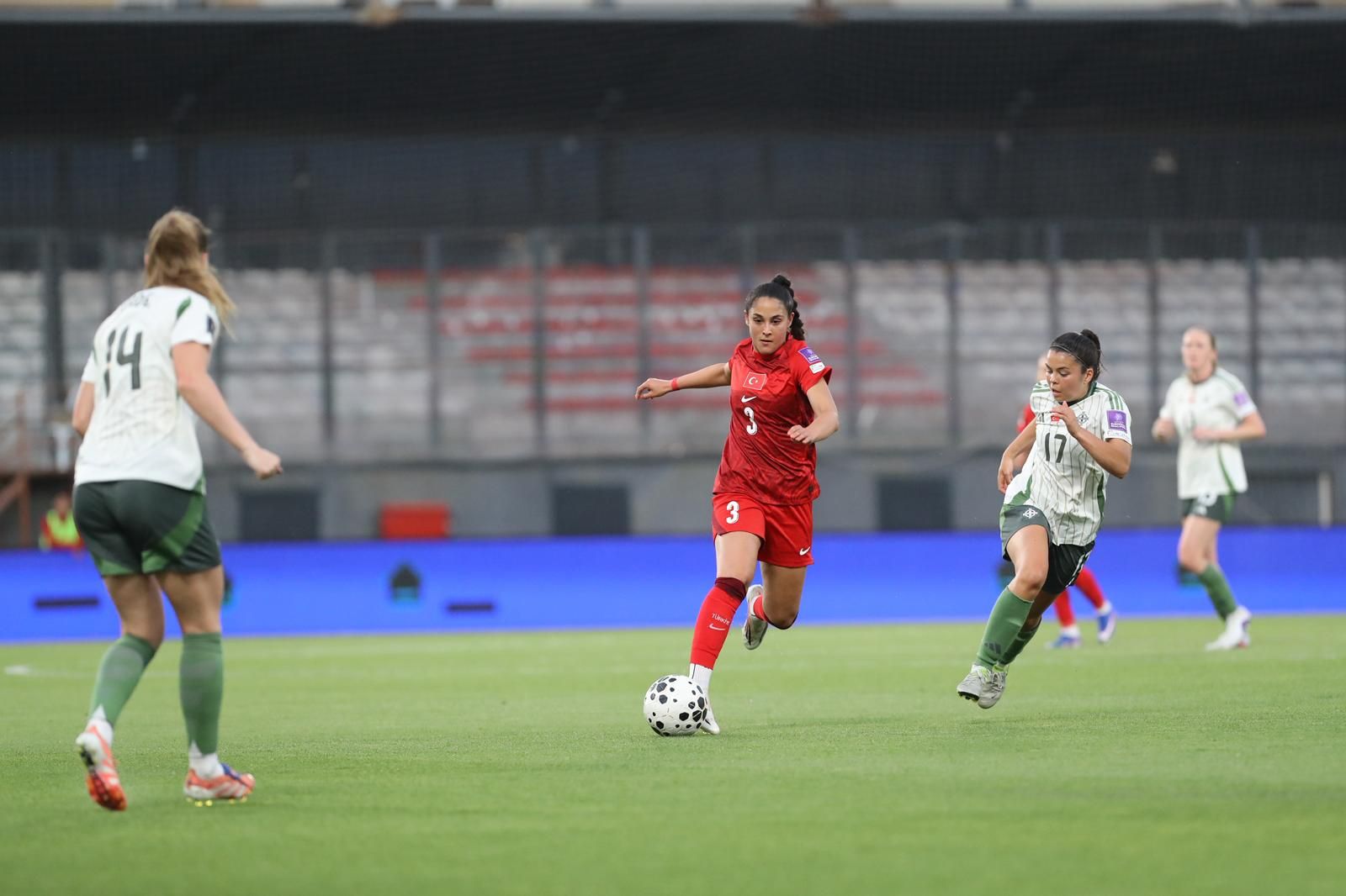 Eine Spielerin in rotem Trikot dribbelt den Ball, während zwei Gegnerinnen in weißen Trikots versuchen, den Ball zu erobern.