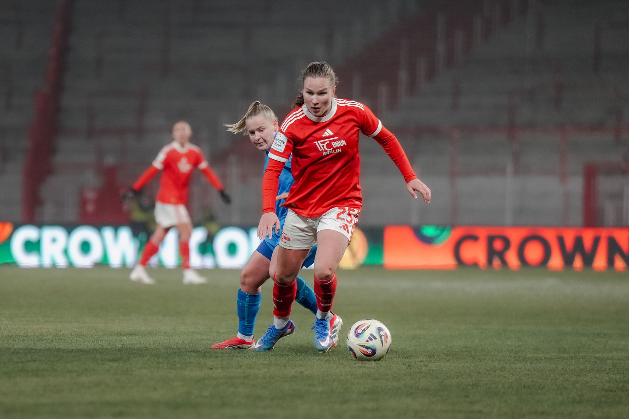 Eine Fußballspielerin in rotem Trikot läuft mit dem Ball, während eine Gegenspielerin in Blau versucht, den Ball zu erobern.
