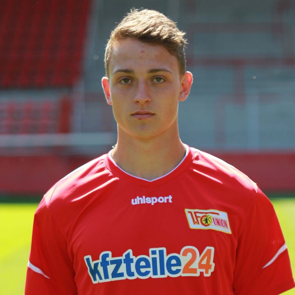 Ein junger Fußballspieler in rotem Trikot steht vor einem Stadionhintergrund und schaut ernst in die Kamera.