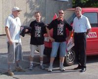 Vier Personen stehen vor einem roten Fahrzeug, einige tragen schwarze T-Shirts mit einem Aufdruck, und es ist sonnig.