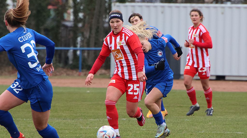 Eine Fußballspielerin in rot-weißer Uniform dribbelt, während eine andere Spielerin in blauer Uniform sie von hinten angreift.