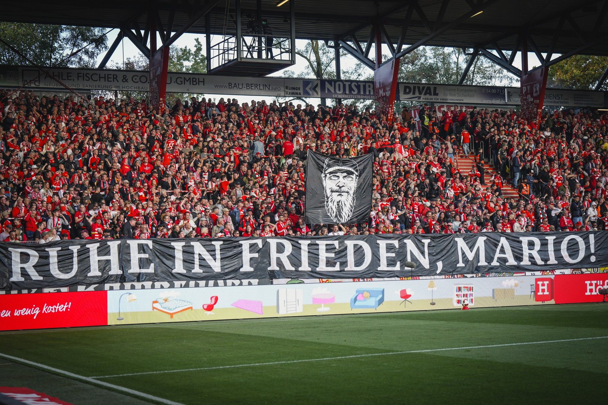 Banner mit der Aufschrift „RUHE IN FRIEDEN, MARIO!“ in einem Stadion, umgeben von zahlreichen Fans in roten Trikots.